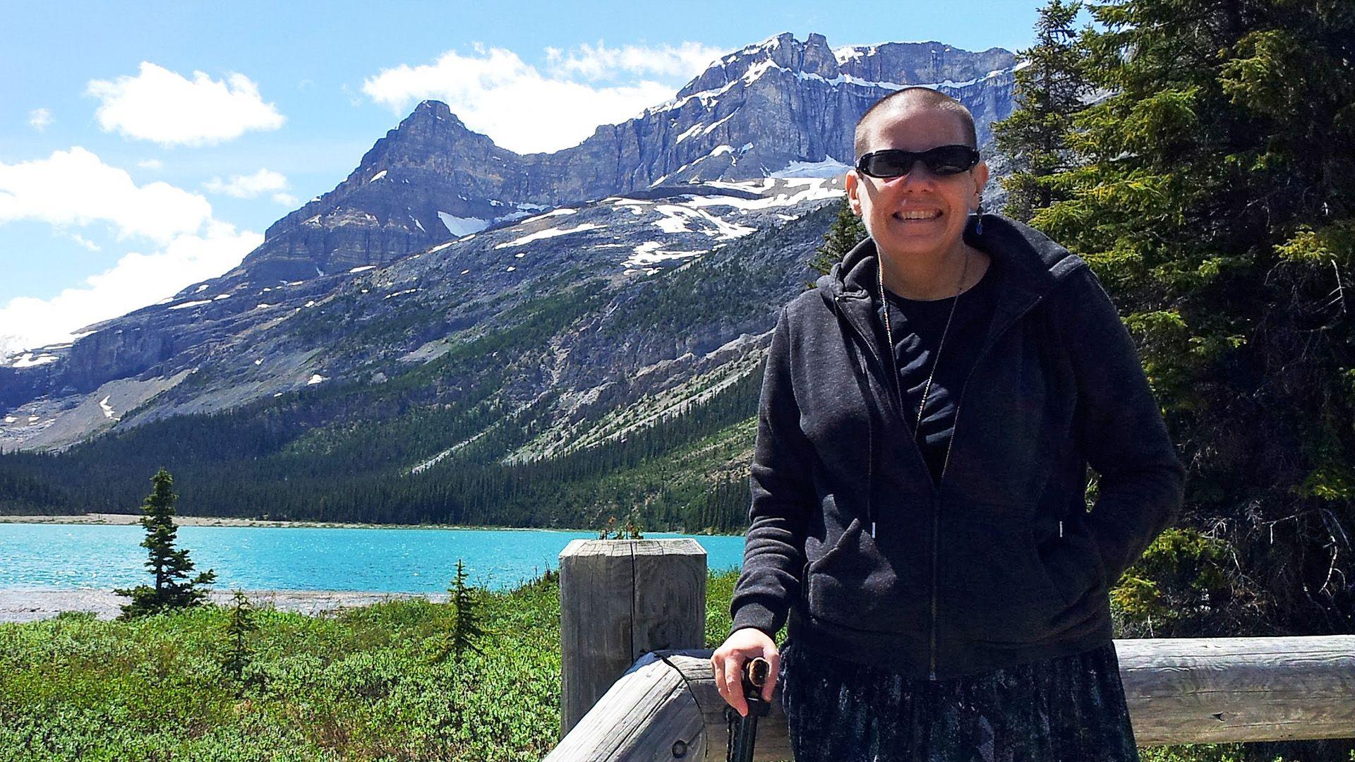 Cindy Baldassi is stood in front of a blue lake and towering mountain peaks with snow visible at highest points
