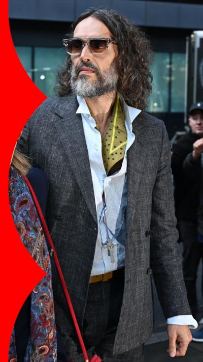 Russell Brand leaving court wearing sunglasses, a medium length grey coat, and a blue shirt with a cravat, with other necklaces. He has long hair and a full salt and pepper beard. 
