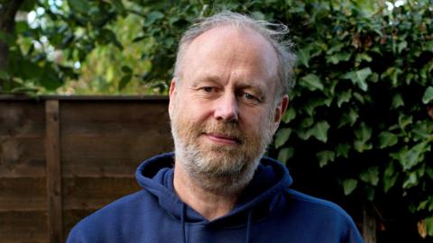 Richard Jackson with a beard and short hair wearing a navy blue hoodie with you are enough written on it in white. He has a neutral expression and there is a fence and foilage behind him.