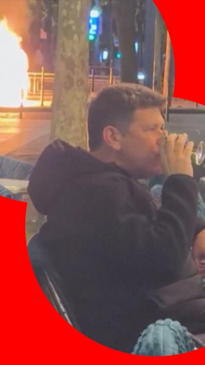 A man sits at the front of a bar in Paris drinking a glass of wine while protests take place behind him 