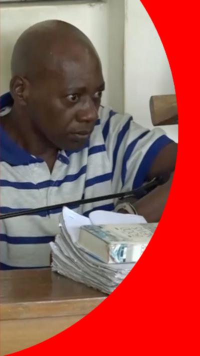 A bald man wearing a white polo-shirt with with a clue collar and blue horizontal stripes. He is sat at a desk on which a white book is placed on top of a pole of white papers.