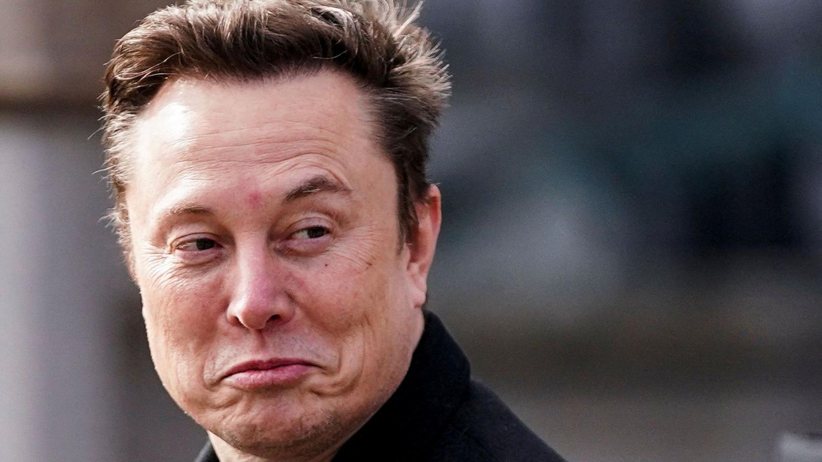 Elon Musk smiles at a camera while appearing to walk by. He is wearing a black coat and has short brown hair