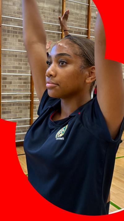 A girl in PE kit has her arms in the air. She is standing in a school gym