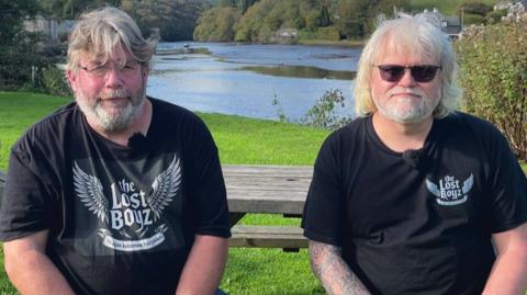 Two men with grey hair sit on a picnic bench with the scenic river in the village of Lerryn behind them. They are wearing black T-shirts bearing the name of their band, Lost Boyz.