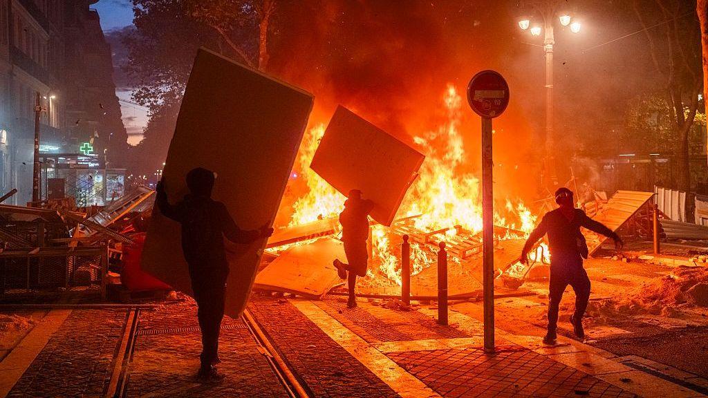 Protesters set barricades on fire in Marseille at night. A large fire can be seen on a city street. Two people are walking towards it holding large rectangular boards, presumably to add to the flames. A third person is walking near the fire. Lampposts, bollards and trees can be seen in the frame, and a row of shops is visible on the left.