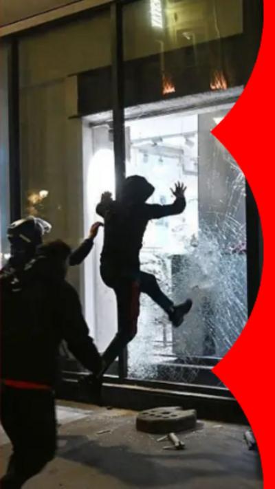 A person wearing a hood, kicking the window of a shop front at night wile others look on.