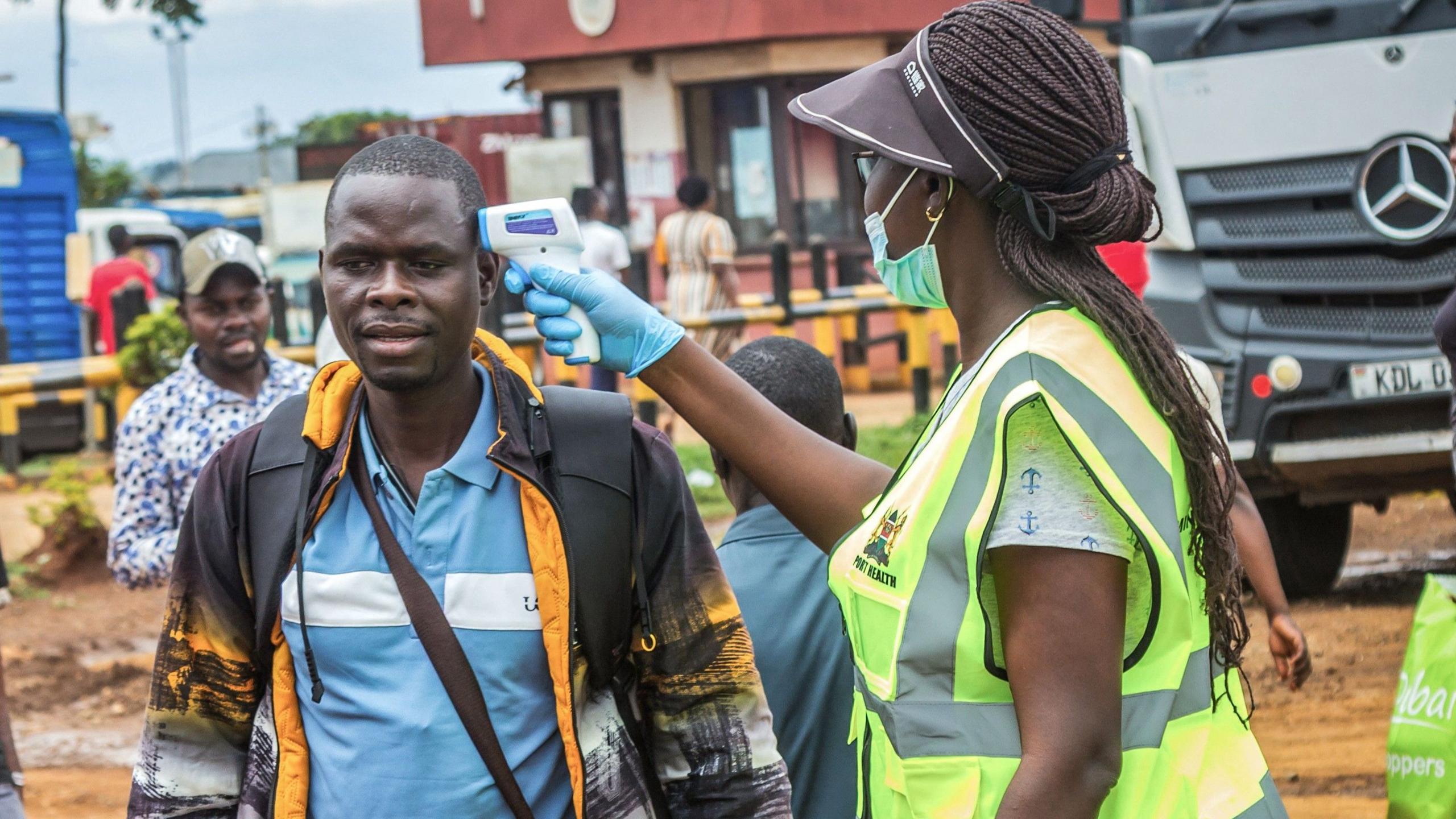 A woman wearing a yellow reflector jacket and a mask checks the temperature of a traveller, a man in a black and yellow jacket and blue shirt 