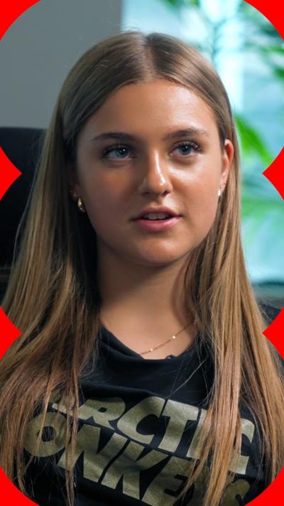 A girl with long blonde hair talks to someone off-camera.
