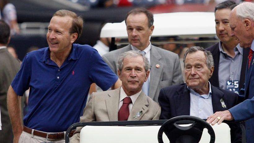 From left: Neil Bush, George W Bush, and George HW Bush. They're sitting in a golf cart and surrounded by security and aides