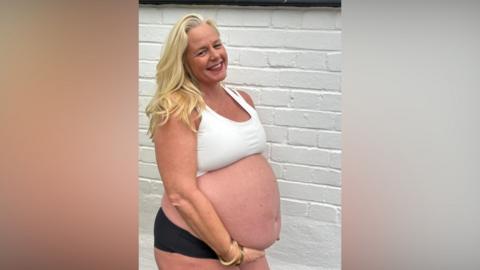 Meghan Mari, a woman in her 50s with blonde hair, holds her pregnant belly. She wears a yoga top and black shorts. 