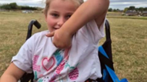 Remi making a thumbs up sign in her wheelchair as she wears a colourful T-shirt with a pink her