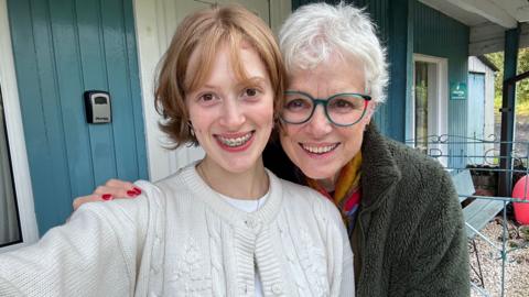 Iona and Judith post for a photo together in front of a wooden cabin with grey-blue doors. Iona is wearing a white knitted cardigan and Judith green glasses and a green fleece.