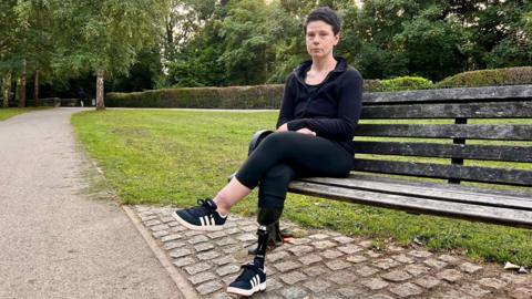A woman in her forties is sitting on a park bench.  She is dressed in black leggings and a sports top.  She has a below knee amputation of her right leg and is missing some of her fingers.