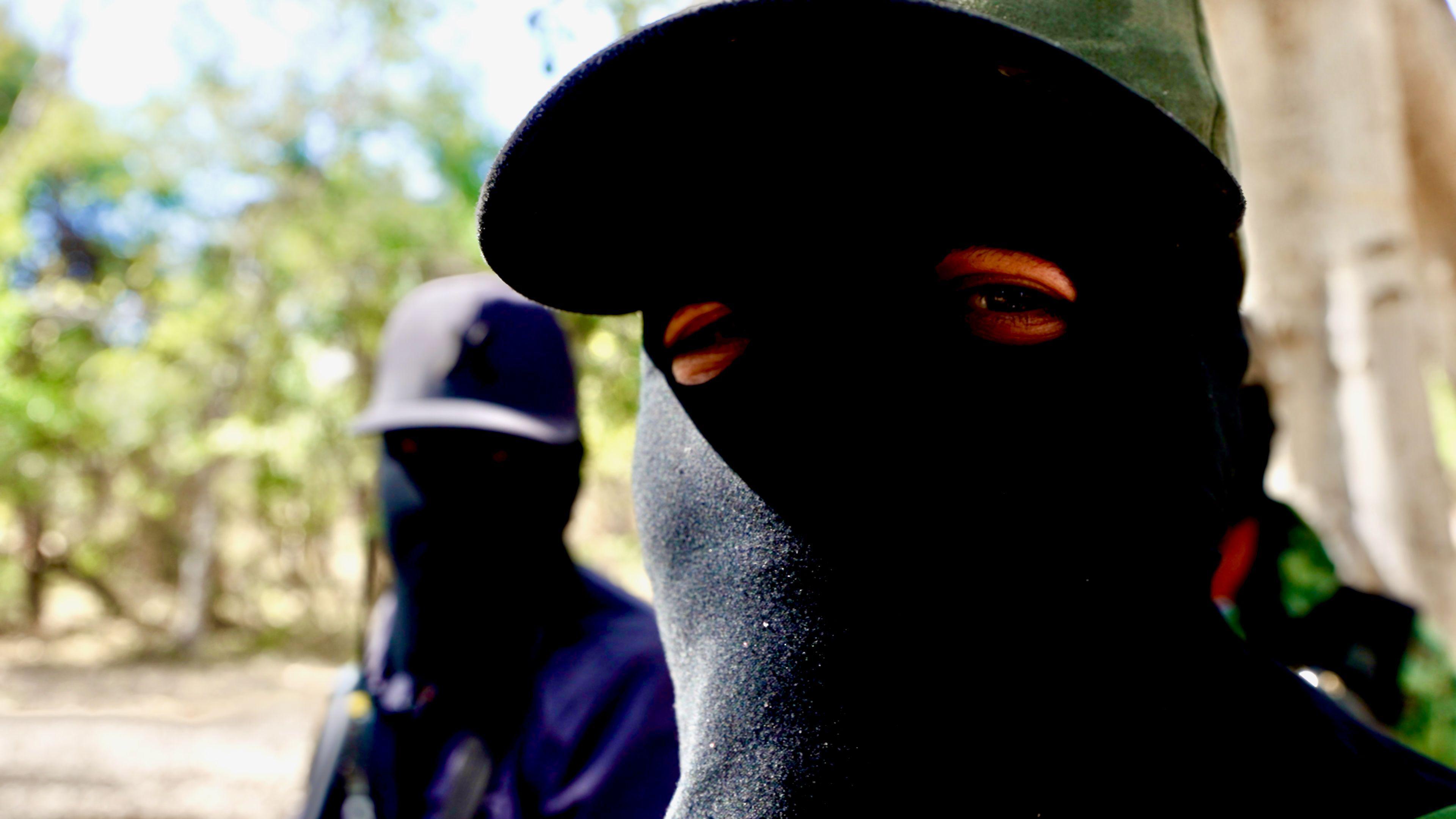 A cartel member wearing a black balaclava looks into the camera, another - also wearing a balaclava, stands in the background.