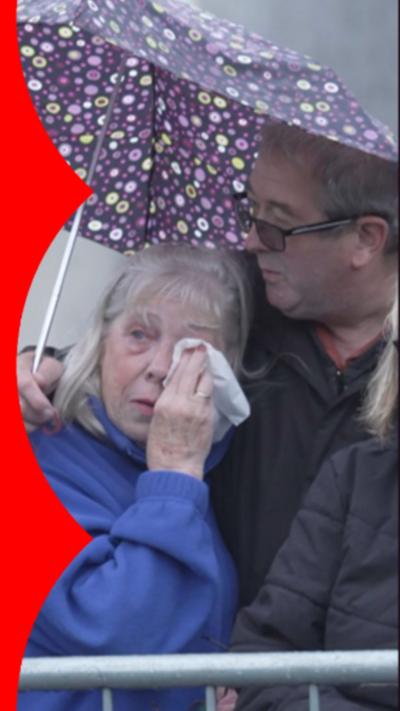 A woman in a blue coat with a handkerchief to her eye. A man is consoling her while holding a purple umbrella