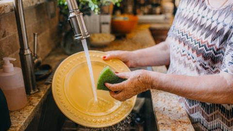 A woman with red nail polish stands over the sink washing a yellow plate. She is wearing a patterned top and can only be seen from her waist to her shoulders.
