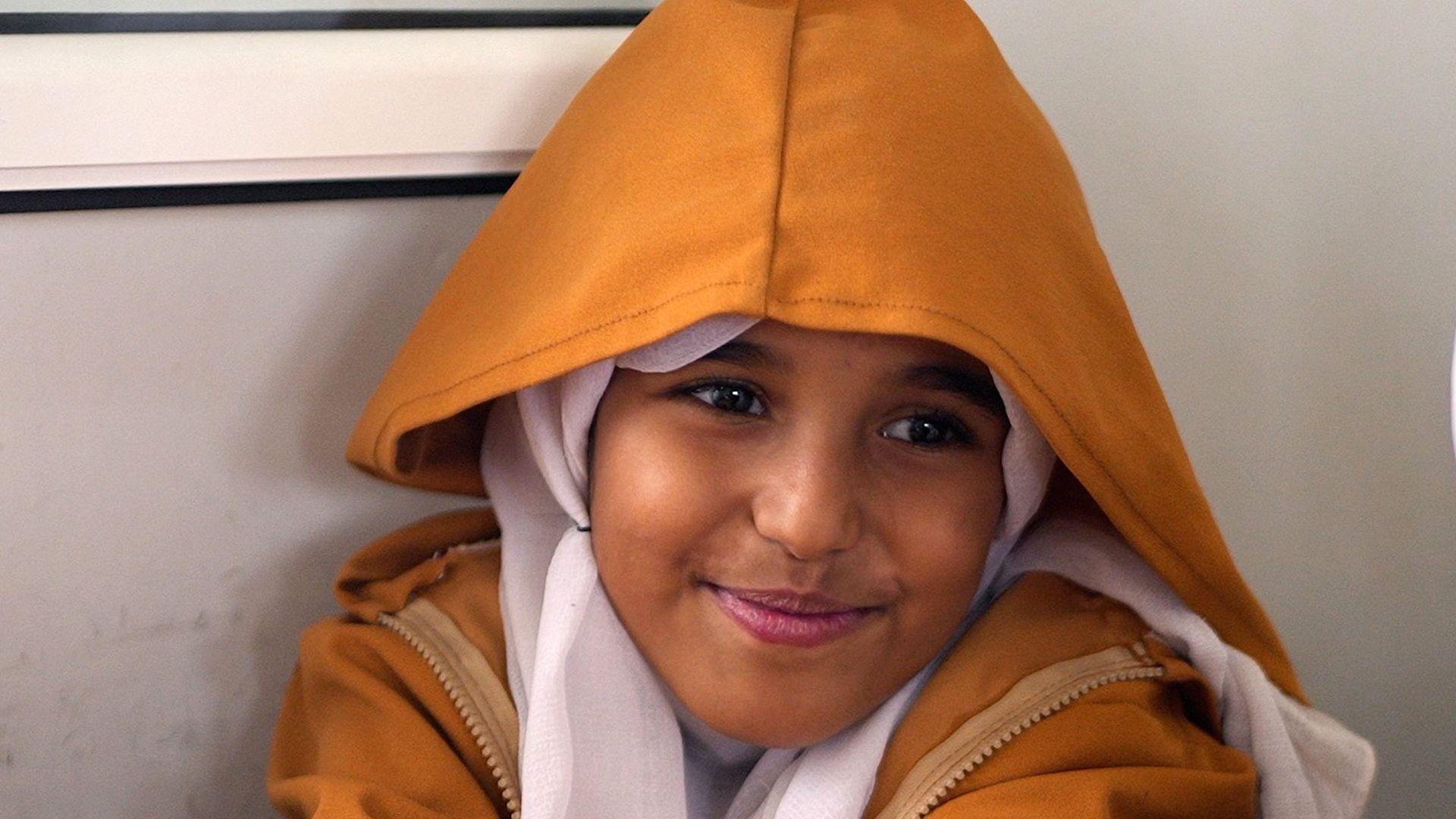 A young girl smiles shyly at the camera. She is wearing a dark yellow hooded coat with a cream headscarf.