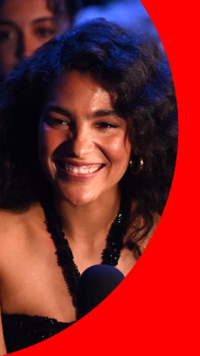 Olivia Dean has long, dark, curly hair and is wearing a black dress.
