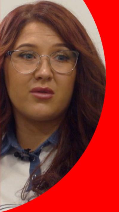 Megan has long, wavy reddish brown hair. She is wearing glasses, a white and blue top, and is smiling at the camera. She is sat on a dark leather chair, with a window and white walls in the background