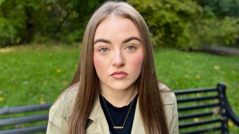 Keeley has a neutral expression. She is wearing a black tshirt, cream trench coay and gold bar necklace with her name on.  She has long hair.