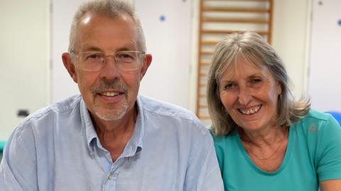 Steve and Angela Bucksey sit side-by-side smiling - Steve has grey hair and is wearing a light blue shirt with a collar, and Angela has shoulder-length greyish hair and wears a green round-necked T-shirt and a necklace