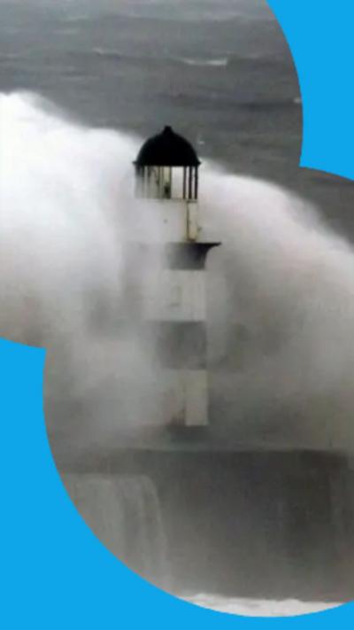 Large waves crashing over a black and white striped lighthouse at the end of a pier.