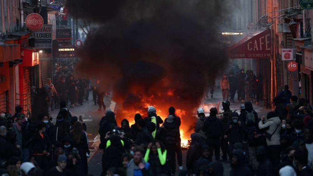 A fire in the middle of a Paris street. Several police offices and demonstrators stand around it.