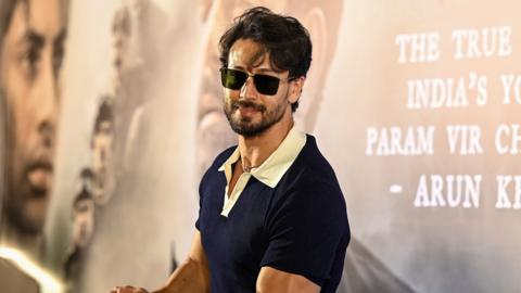 Tiger Shroff poses at a promotional event, wearing dark sunglasses and a navy polo shirt with a white collar. The actor leans on a display stand in front of a backdrop featuring large portrait images and text from a film campaign.
