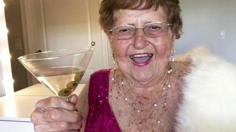 Ninety-six-year-old Grandma Droniak, holding a martini glass and wearing a pink sparkly top and a white fur shawl. She is standing in front of a dressing table with lights around the mirror.