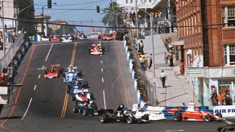 Formula 1 cars head down Linden Avenue during the 1976 United States Grand Prix West at Long Beach. Ferrari's Niki Lauda leads the pack while among those following him are Ronnie Peterson, Tom Pryce, Jean-Pierre Jarier, John Watson, Jody Scheckter, Jacques Laffite, Carlos Pace, Jochen Mass, Mario Andretti, Emerson Fittipaldi and Chris Amon