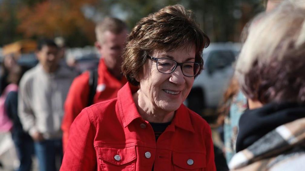 Susan Collins in a red jacket and glasses in a crowd of people