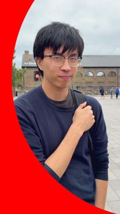 A man with short black hair and glasses stands in an outdoor piazza