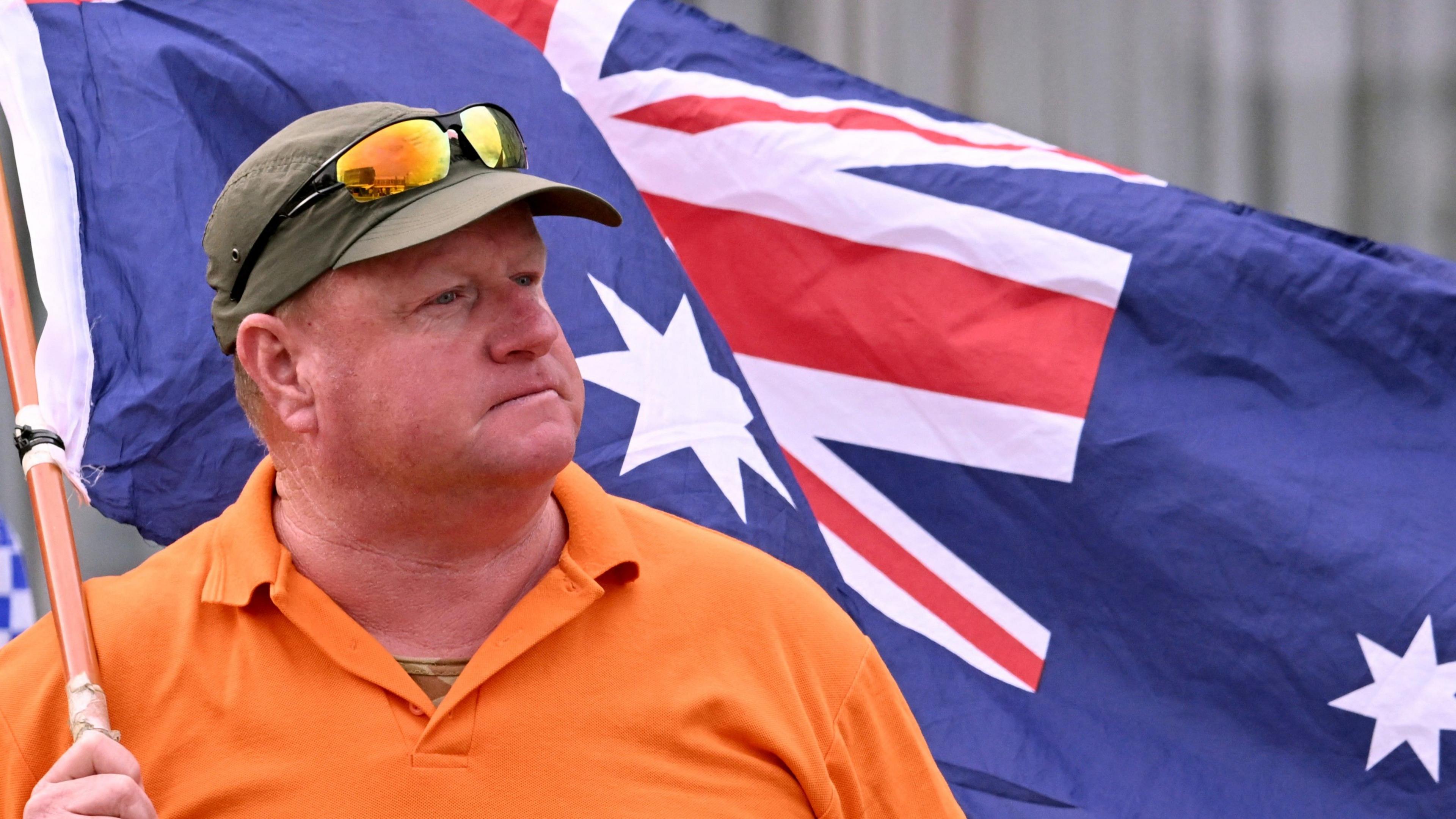 A man wearing an orange t-shirt reading "Wangaratta this is not us!!" waves an Australian flag