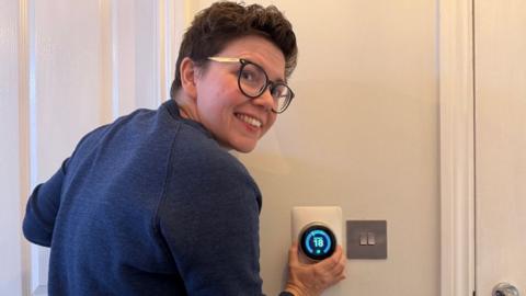 Cora Harrison, a woman with short brown hair and glasses in a navy jumper, adjusts the temperature on a thermostat to 18 degrees