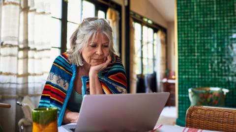 Old lady with grey hair sits at home with a brightly coloured blanket around her shoulders, looking at her laptop