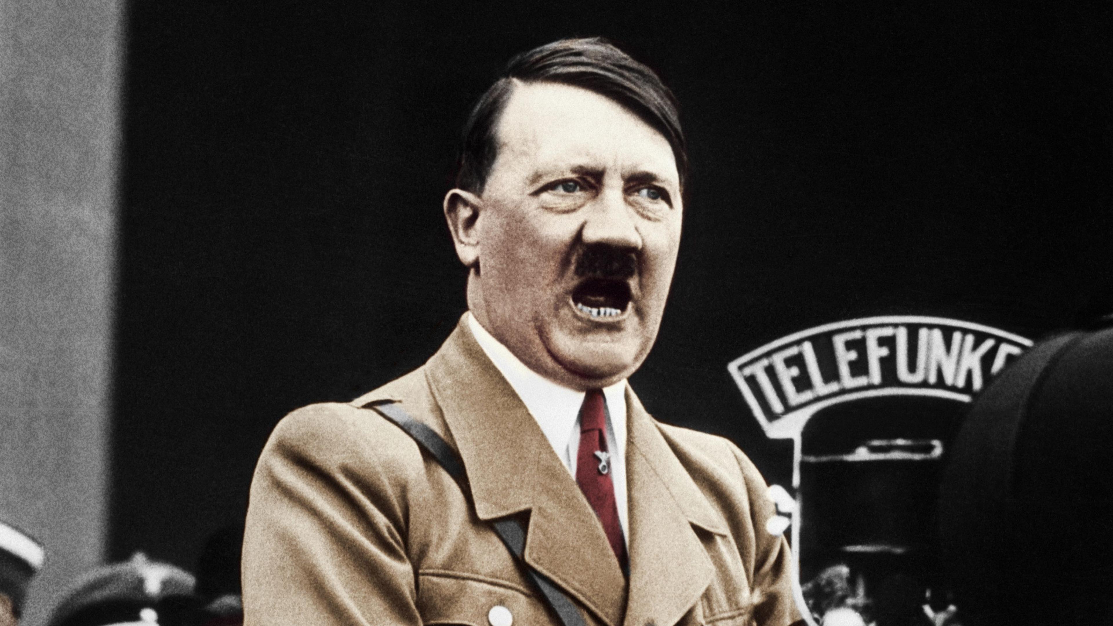 A colour treated photo of Hitler speaking into a big microphone. He's wearing a light brown jacket and red tie.