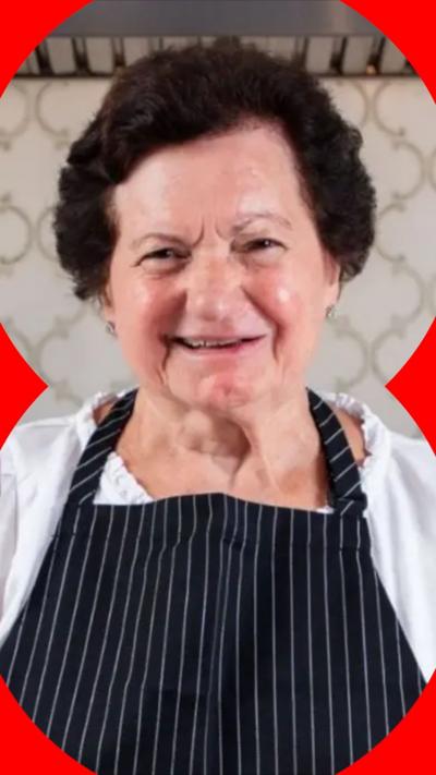 An elderly woman with dark hair smiles wearing a blue pinstripe apron over a white top. She holds a glass of red wine