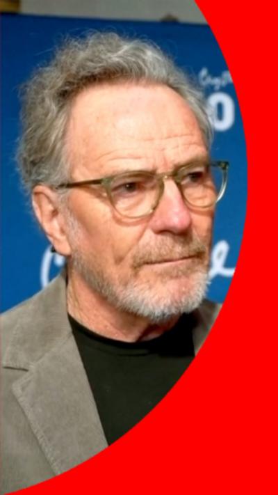 Bryan Cranston has short light hair and short grey bread. He is wearing glasses, and a grey blazer over a black shirt.