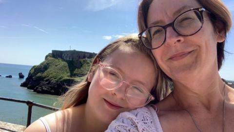 A young girls wearing glasses with blonde hair rests her chin on the shoulder of a woman with brown hair and glasses. An island surrounded by water is seen behind them.